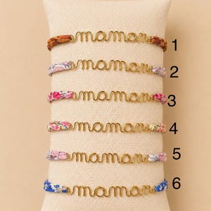 Op de afbeelding: Zes goudkleurige draadarmbanden met de tekst "mama" worden getoond op een beige kussen. De armbanden hebben verschillende bandontwerpen, waaronder gevlochten bruin, bloempatronen in roze en blauw, en een blauw bloemmotief.
