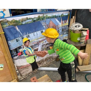 May include: Two children wearing yellow hard hats play with a red brick in front of a backdrop of a building site. The backdrop shows a row of new houses under construction.