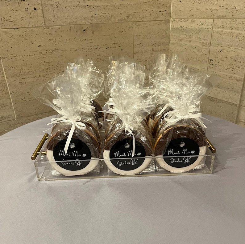 May include: Three round, black and white dipped cookies individually wrapped in clear cellophane bags with white ribbon ties. Each cookie features a circular black label with the white text "Meet Me @ Studio BK" displayed on a clear tray.