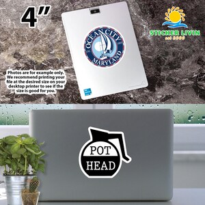 May include: A white sticker with a black graphic of a coffee pot silhouette and the text POT HEAD in black letters, applied to a gray laptop. A second sticker featuring an Ocean City Maryland sailboat logo is visible on a white tablet above.