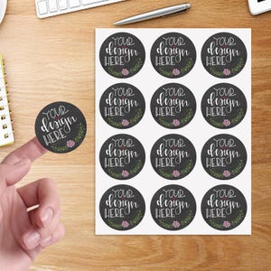 May include: A sheet of twelve circular stickers and one individual sticker featuring a dark gray background with white cursive text that reads YOUR DESIGN HERE, accented by a small pink flower and green leafy vine graphic at the bottom.