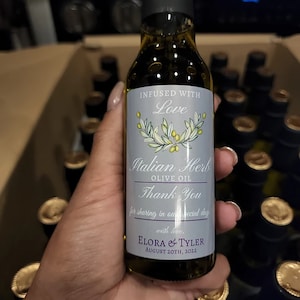 May include: A glass bottle of Italian Herb olive oil with a light blue label featuring a green olive branch illustration and text that reads Infused with Love, Italian Herb Olive Oil, Thank You for sharing in our special day, Elora & Tyler, August 20th, 2022.