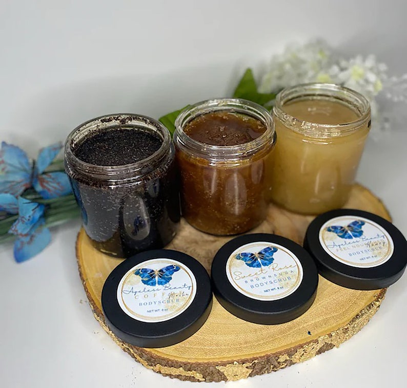 May include: Three clear plastic jars filled with body scrub, each topped with a black lid featuring a butterfly logo and the text Ageless Beauty. The jars contain dark coffee, brown sugar, and light honey-colored scrubs, each labeled 8 oz.