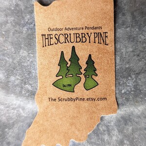 May include: A brown leather-like tag shaped like the state of Indiana with the text "Outdoor Adventure Pendants THE SCRUBBY PINE Est. 1994 The ScrubbyPine.etsy.com" printed on it. The tag features a green silhouette of three pine trees.