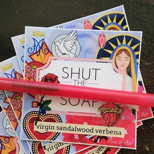 May include: A stack of colourful soap bars with a religious theme. The soap bars are decorated with images of a dove, a heart, and a woman with a crown. The text "SHUT THE SOAP" is printed on the top of the bars. The soap bars are labelled with the scents "virgin sandalwood verbena" and "Haus of Aza".