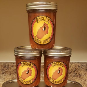 May include: Three glass jars of Rocco's Secret Gravy, stacked on top of each other. The jars have a red, yellow, and orange label with the text "Rocco's Secret Gravy" and a cartoon hand.