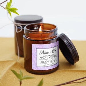 May include: A lit candle in an amber glass jar with a black lid and a label reading Amore Co. HAND MADE WAX CANDLES BLOSSOM NET WT. 2.75 OZ. A second identical candle jar with a lid and twine sits behind it, accompanied by green leafy branches.