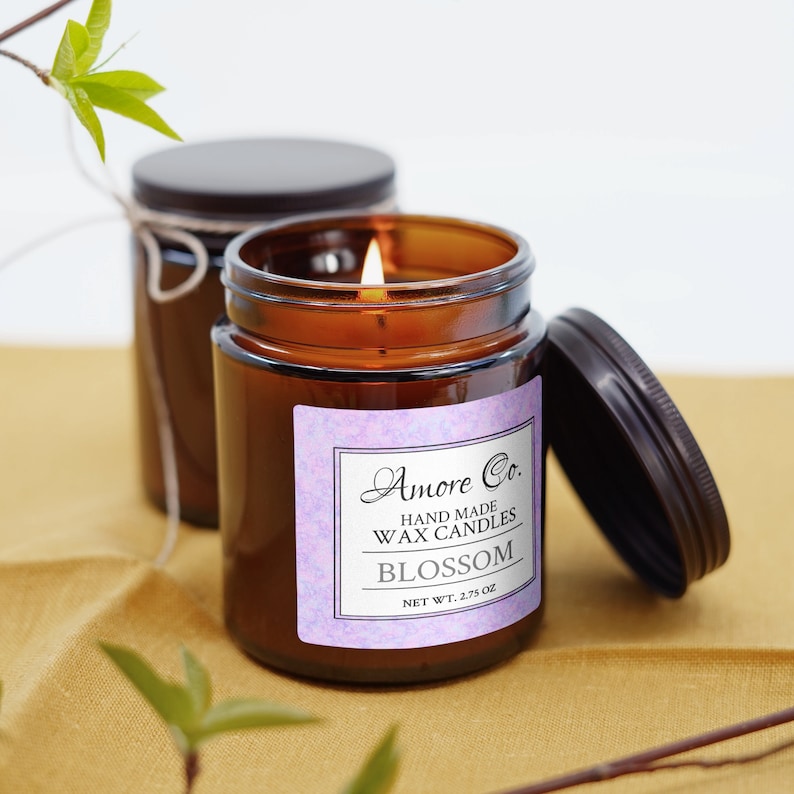 May include: A lit candle in an amber glass jar with a black lid and a label reading Amore Co. HAND MADE WAX CANDLES BLOSSOM NET WT. 2.75 OZ. A second identical candle jar with a lid and twine sits behind it, accompanied by green leafy branches.