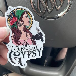May include: A white sticker featuring a colourful illustration of a woman with a green headscarf and a tattoo machine. The text "The Tattooed Gypsy" is written in black below the illustration.