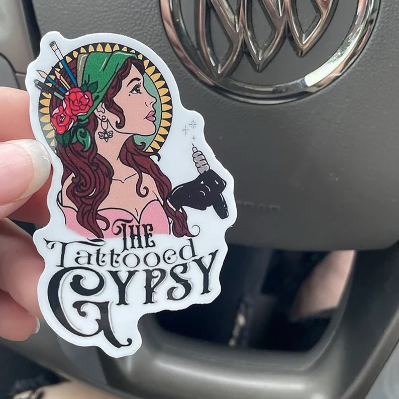 May include: A white sticker featuring a colourful illustration of a woman with a green headscarf and a tattoo machine. The text "The Tattooed Gypsy" is written in black below the illustration.
