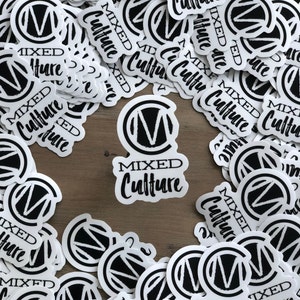 May include: A collection of white stickers with a black and white design. Each sticker features the words "MIXED Culture" in a stylized font, with a circular logo containing the letter "M". The stickers are scattered on a wooden surface.