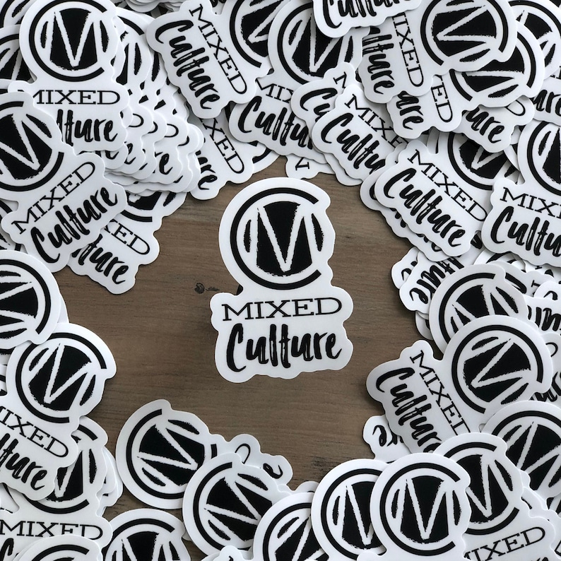 May include: A collection of white stickers with a black and white design. Each sticker features the words "MIXED Culture" in a stylized font, with a circular logo containing the letter "M". The stickers are scattered on a wooden surface.