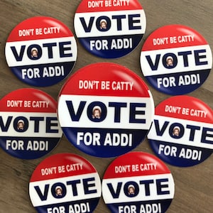 May include: A collection of round, red, white, and blue buttons. Each button has the text "DON'T BE CATTY" at the top, "VOTE" in the center, and "FOR ADDI" at the bottom. A small image of a person is in the center of the "VOTE" text.