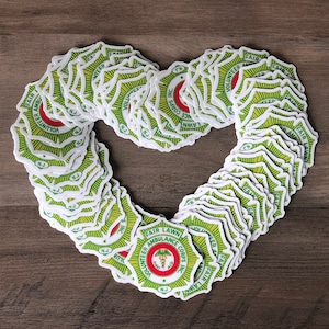 May include: A collection of stickers arranged in a heart shape. Each sticker features a green and yellow design with the words "FAIR LAWN AMBULANCE CORPS INC." and a medical symbol. The stickers are on a wooden surface.