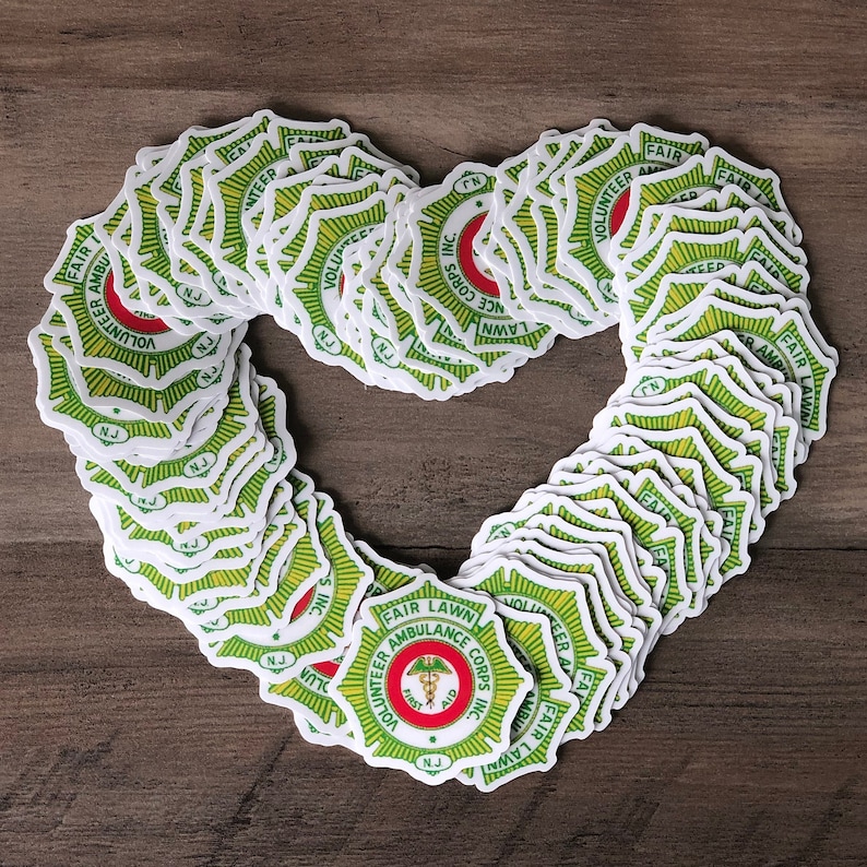 May include: A collection of stickers arranged in a heart shape. Each sticker features a green and yellow design with the words "FAIR LAWN AMBULANCE CORPS INC." and a medical symbol. The stickers are on a wooden surface.
