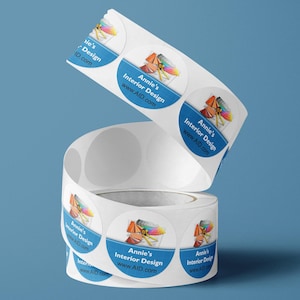 May include: A roll of circular white adhesive labels featuring a blue horizontal band with the text Annie's Interior Design and www.AID.com. Above the text is a graphic illustration of a yellow measuring tape and a colorful paint fan deck.