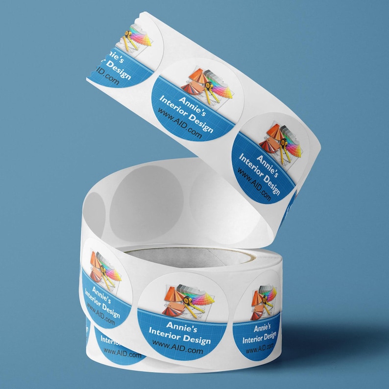 May include: A roll of circular white adhesive labels featuring a blue horizontal band with the text Annie's Interior Design and www.AID.com. Above the text is a graphic illustration of a yellow measuring tape and a colorful paint fan deck.