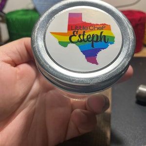 May include: A silver jar lid with a white sticker featuring a rainbow-coloured outline of the state of Texas. The sticker also includes the text "Little Chef Esteph".