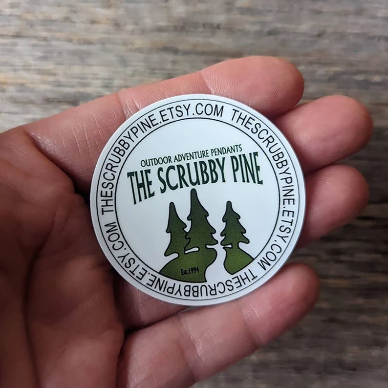 May include: A round white sticker with the text "THE SCRUBBY PINE" and three green pine trees. The sticker also includes the text "Outdoor Adventure Pendants" and "EST. 1994". The website address thescrubbypine.etsy.com is printed around the edge.
