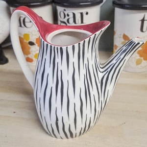 May include: A white ceramic jug with a black zebra stripe design. The rim is red, and it has a white handle. The jug is a decorative item, possibly for serving drinks.