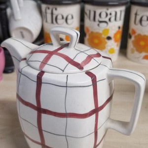 May include: White ceramic teapot with a red and grey plaid design. The teapot features a curved spout, a handle, and a lid with a small handle. Background includes coffee, sugar, and tea canisters.