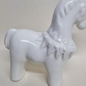 May include: A small, white ceramic horse figurine. The horse has a stylized design with a curved neck and a detailed mane. The figurine is standing on four legs and has a small tail. The horse is a decorative item.