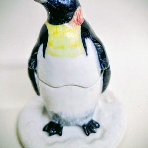 May include: A ceramic penguin ornament with a black head, back, and flippers, a yellow chest, and a white belly. The penguin stands on a white, round base. The beak is white and the neck has red accents. The figurine is a decorative item.