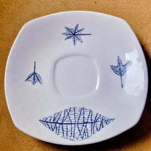 May include: A white ceramic saucer with a square shape and rounded corners. The saucer features blue leaf and starburst designs. The centre has a recessed area for a cup. The saucer is likely part of a tea or coffee set.