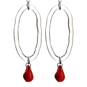 May include: A pair of clear, oval-shaped earrings with a textured surface. Each earring features a small, bright red teardrop-shaped charm suspended from a silver-colored metal loop. The earrings are set against a white background.