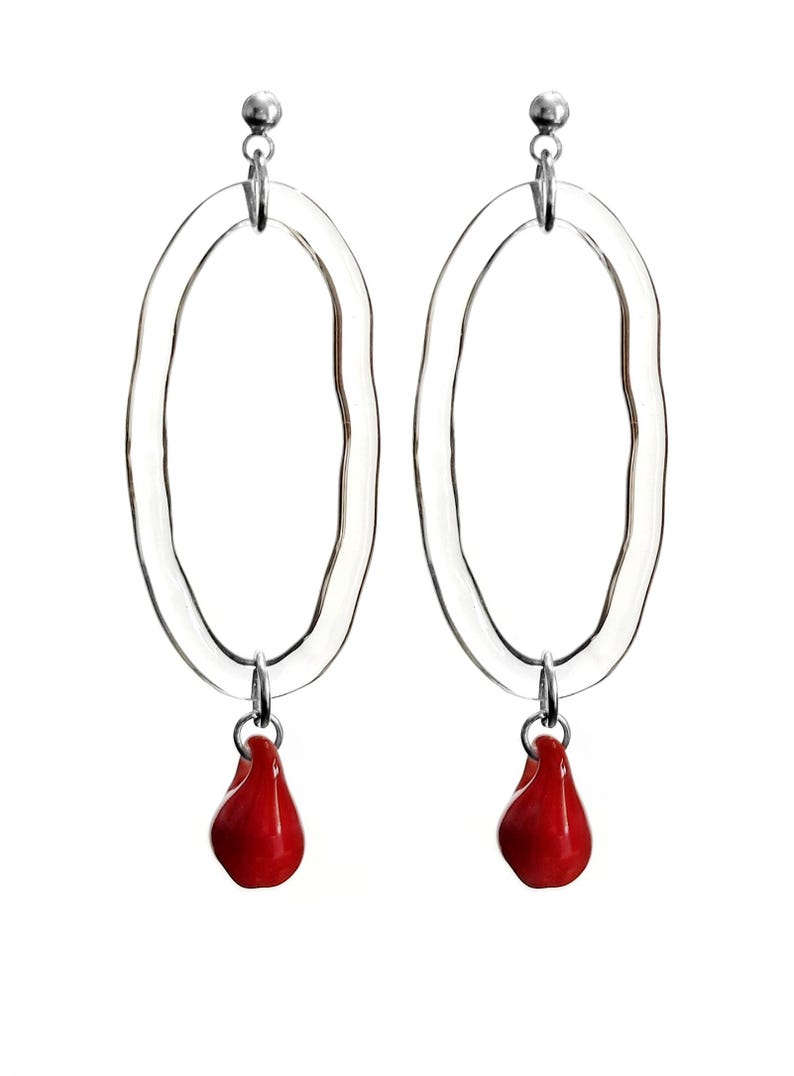 May include: A pair of clear, oval-shaped earrings with a textured surface. Each earring features a small, bright red teardrop-shaped charm suspended from a silver-colored metal loop. The earrings are set against a white background.