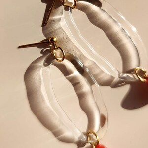 May include: Clear acrylic hoop earrings with gold-colored accents and a red teardrop-shaped charm. The earrings have a post-back closure and are displayed on a light-colored surface.