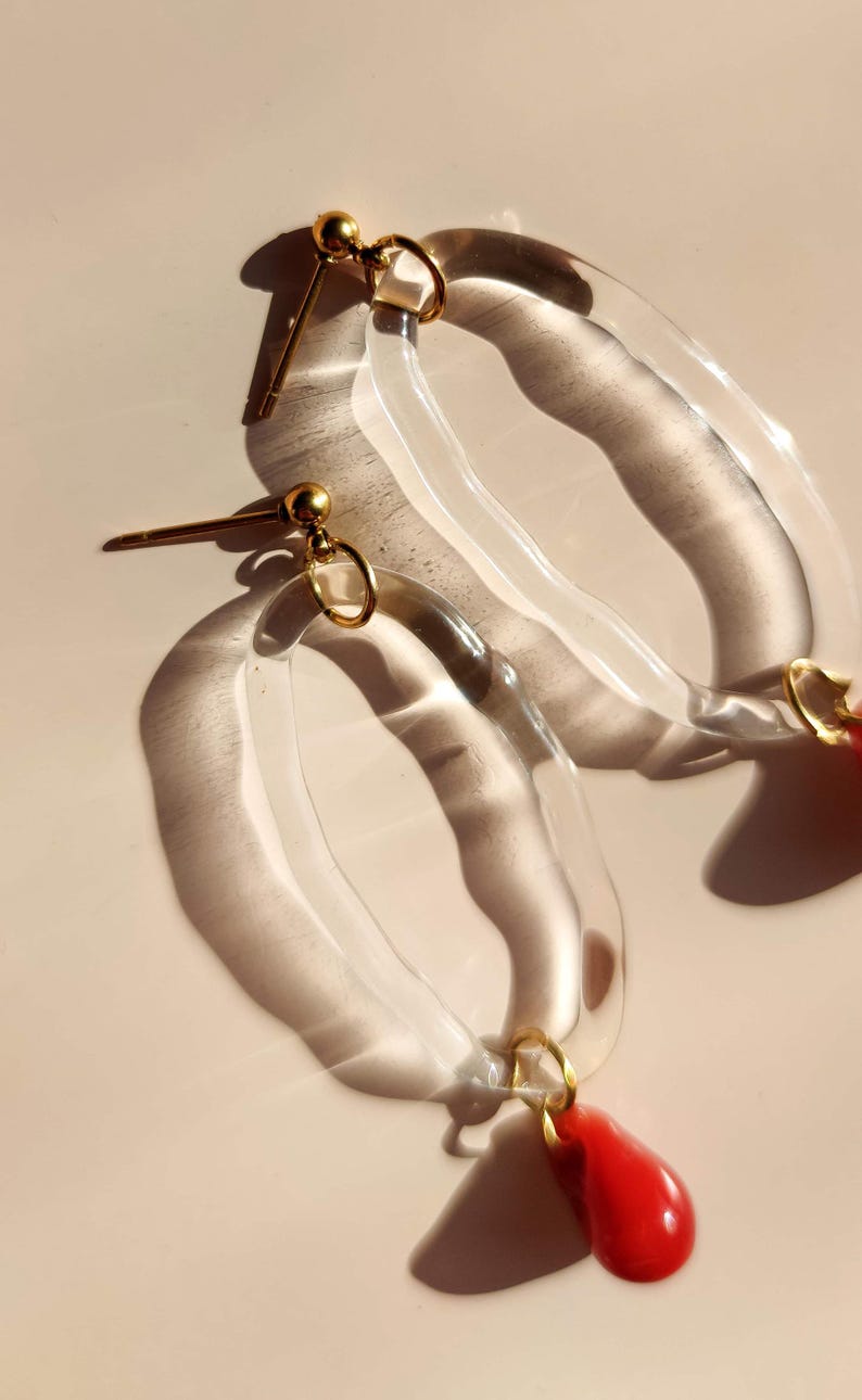 May include: Clear acrylic hoop earrings with gold-colored accents and a red teardrop-shaped charm. The earrings have a post-back closure and are displayed on a light-colored surface.