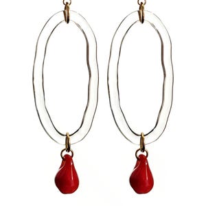 May include: A pair of clear oval earrings with a wavy texture. Each earring has a gold-colored stud and a small, red teardrop-shaped charm hanging from the bottom. The earrings are set against a white background.