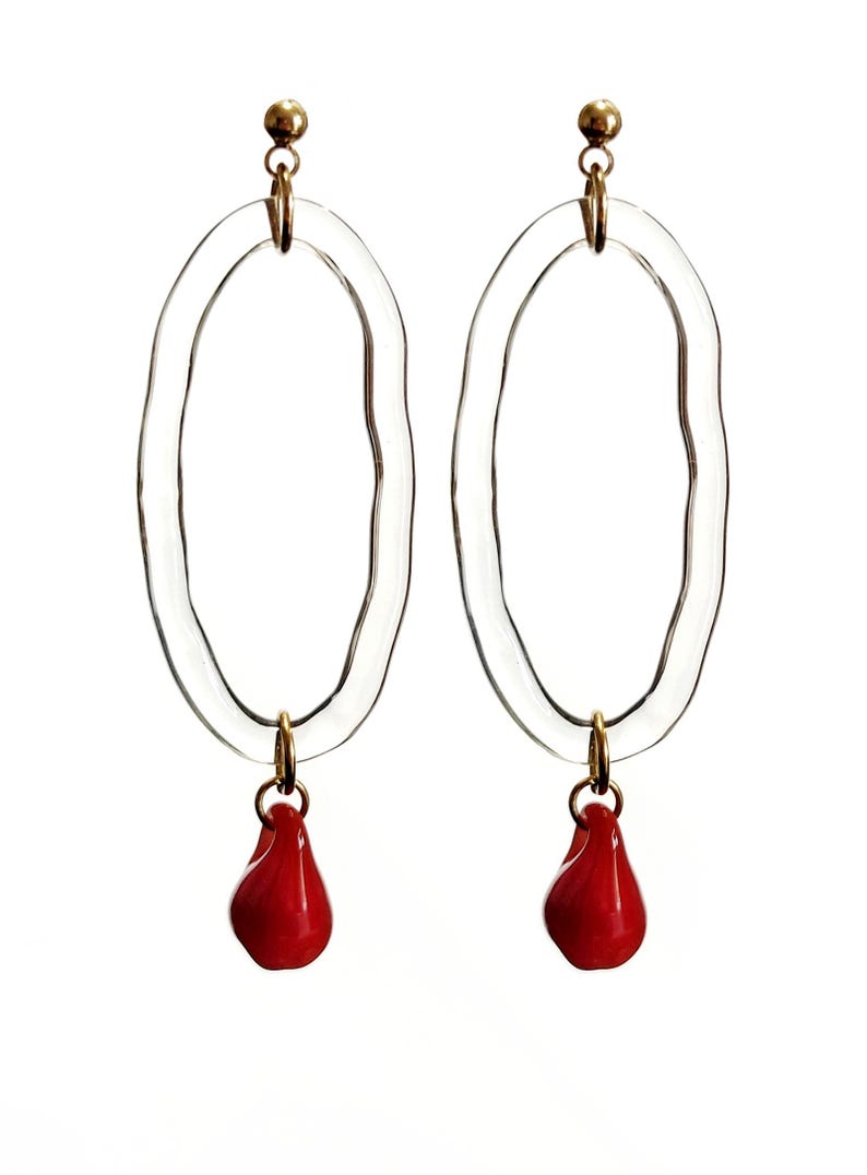 May include: A pair of clear oval earrings with a wavy texture. Each earring has a gold-colored stud and a small, red teardrop-shaped charm hanging from the bottom. The earrings are set against a white background.