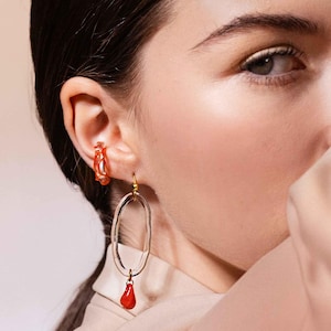 May include: A pair of earrings featuring a clear, oval-shaped design with a small, red teardrop pendant. The earrings are worn on a person's ear, with a matching orange hoop earring visible. The earrings have a gold-colored hook.