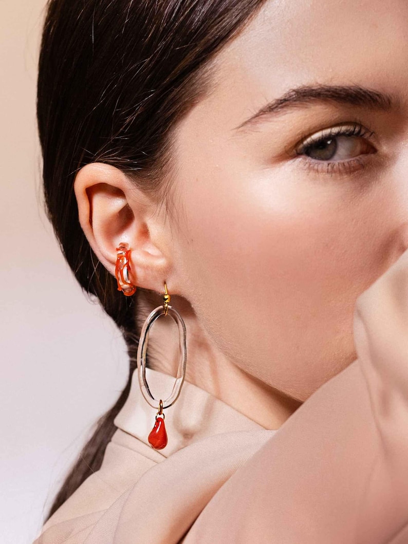 May include: A pair of earrings featuring a clear, oval-shaped design with a small, red teardrop pendant. The earrings are worn on a person's ear, with a matching orange hoop earring visible. The earrings have a gold-colored hook.