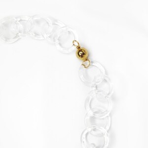 Pode incluir: Um colar de corrente de pl&aacute;stico transparente com um detalhe de contas douradas. A corrente &eacute; feita de c&iacute;rculos grandes e interligados.