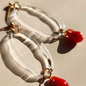 May include: Clear, oval-shaped earrings with a gold-colored post and ring, each featuring a vibrant red, teardrop-shaped charm. The earrings are displayed on a light beige surface, with shadows adding depth.