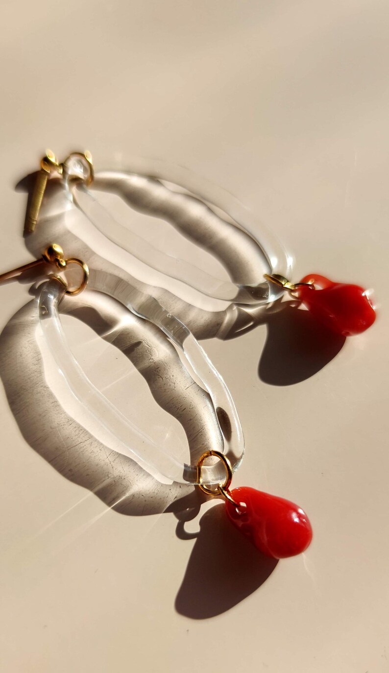 May include: Clear, oval-shaped earrings with a gold-colored post and ring, each featuring a vibrant red, teardrop-shaped charm. The earrings are displayed on a light beige surface, with shadows adding depth.