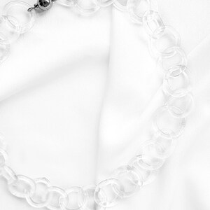 Pode incluir: Um colar de corrente de pl&aacute;stico transparente com um fecho prateado. O colar est&aacute; sobre um fundo de tecido branco.