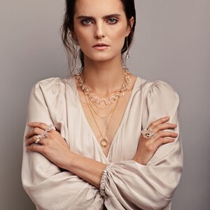 Pode incluir: Uma mulher vestindo uma blusa bege claro com um colar de ouro em camadas e um colar de cristal transparente. Ela est&aacute; usando um anel de cristal transparente na m&atilde;o esquerda e uma pulseira de cristal transparente no pulso direito.