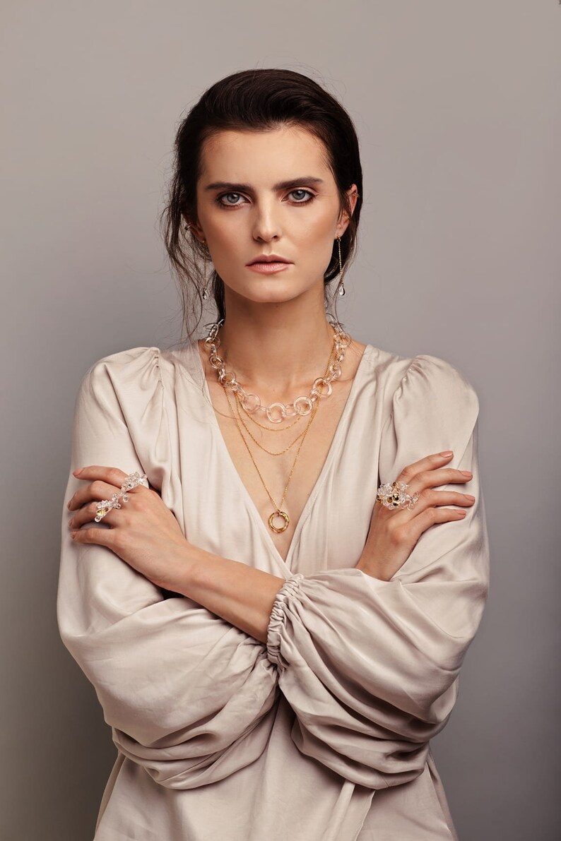 Pode incluir: Uma mulher vestindo uma blusa bege claro com um colar de ouro em camadas e um colar de cristal transparente. Ela est&aacute; usando um anel de cristal transparente na m&atilde;o esquerda e uma pulseira de cristal transparente no pulso direito.