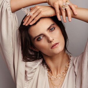 Pode incluir: Uma mulher vestindo uma blusa bege com um colar em camadas que inclui uma corrente de cristal transparente, uma corrente de ouro com um pingente circular e uma corrente de ouro delicada. Ela tamb&eacute;m est&aacute; usando um par de brincos de cristal transparente e dois an&eacute;is de cristal.