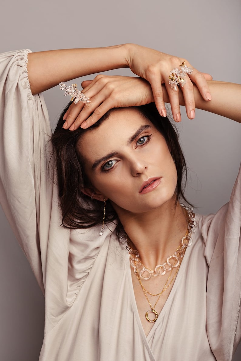 Pode incluir: Uma mulher vestindo uma blusa bege com um colar em camadas que inclui uma corrente de cristal transparente, uma corrente de ouro com um pingente circular e uma corrente de ouro delicada. Ela tamb&eacute;m est&aacute; usando um par de brincos de cristal transparente e dois an&eacute;is de cristal.