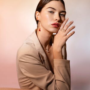 May include: A woman wearing gold jewelry, including earrings with red accents, a necklace, and a large ring. She is wearing a beige blazer. The background has a gradient of pink and white.