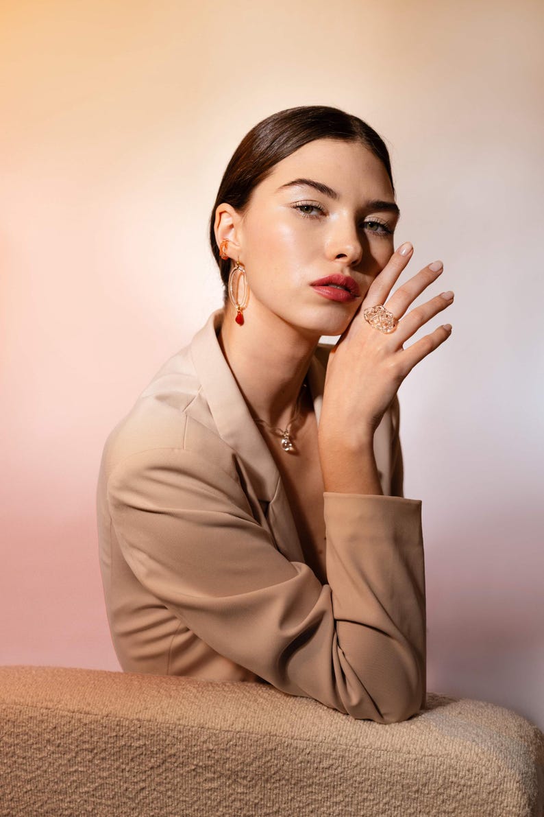 May include: A woman wearing gold jewelry, including earrings with red accents, a necklace, and a large ring. She is wearing a beige blazer. The background has a gradient of pink and white.