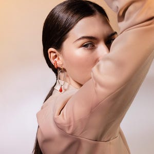 May include: A woman wearing gold-toned earrings with red accents. One earring is a small hoop, and the other is an oval shape with a dangling red teardrop. The woman is wearing a tan blazer and has dark hair.