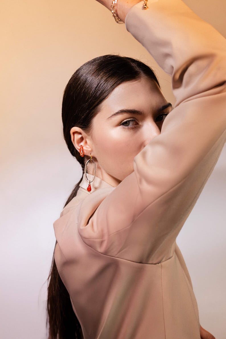 May include: A woman wearing gold-toned earrings with red accents. One earring is a small hoop, and the other is an oval shape with a dangling red teardrop. The woman is wearing a tan blazer and has dark hair.
