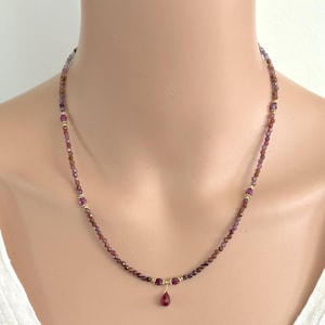 May include: A delicate necklace featuring small, faceted, reddish-purple beads. The necklace has gold-colored accents and a teardrop-shaped pendant. The necklace is displayed on a person's neck.