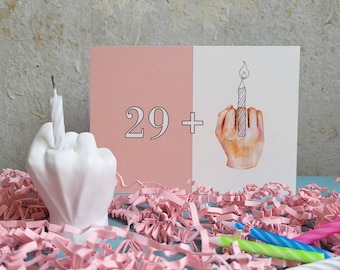 May include: A rectangular greeting card featuring a split pink and white background with the text 29 + and an illustration of a hand making a middle finger gesture holding a lit candle. A white hand-shaped candle holder holds a single white candle.